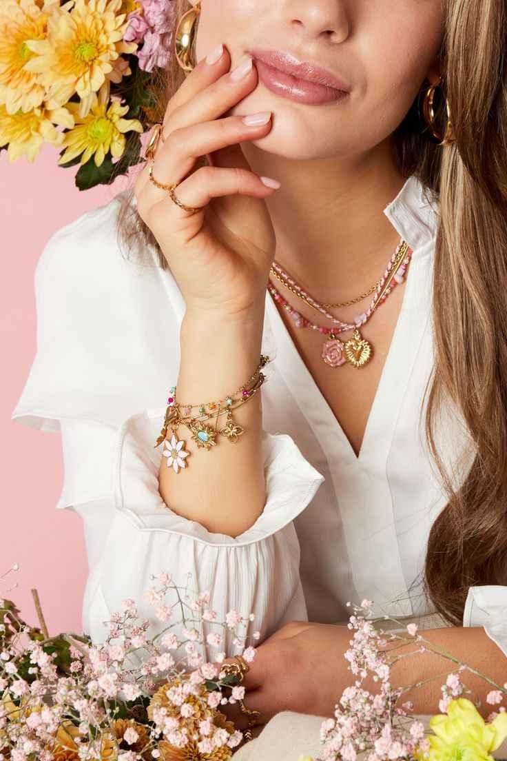 Close-up of woman in white blouse wearing gold charm bracelet and layered necklaces with flowers on pink background