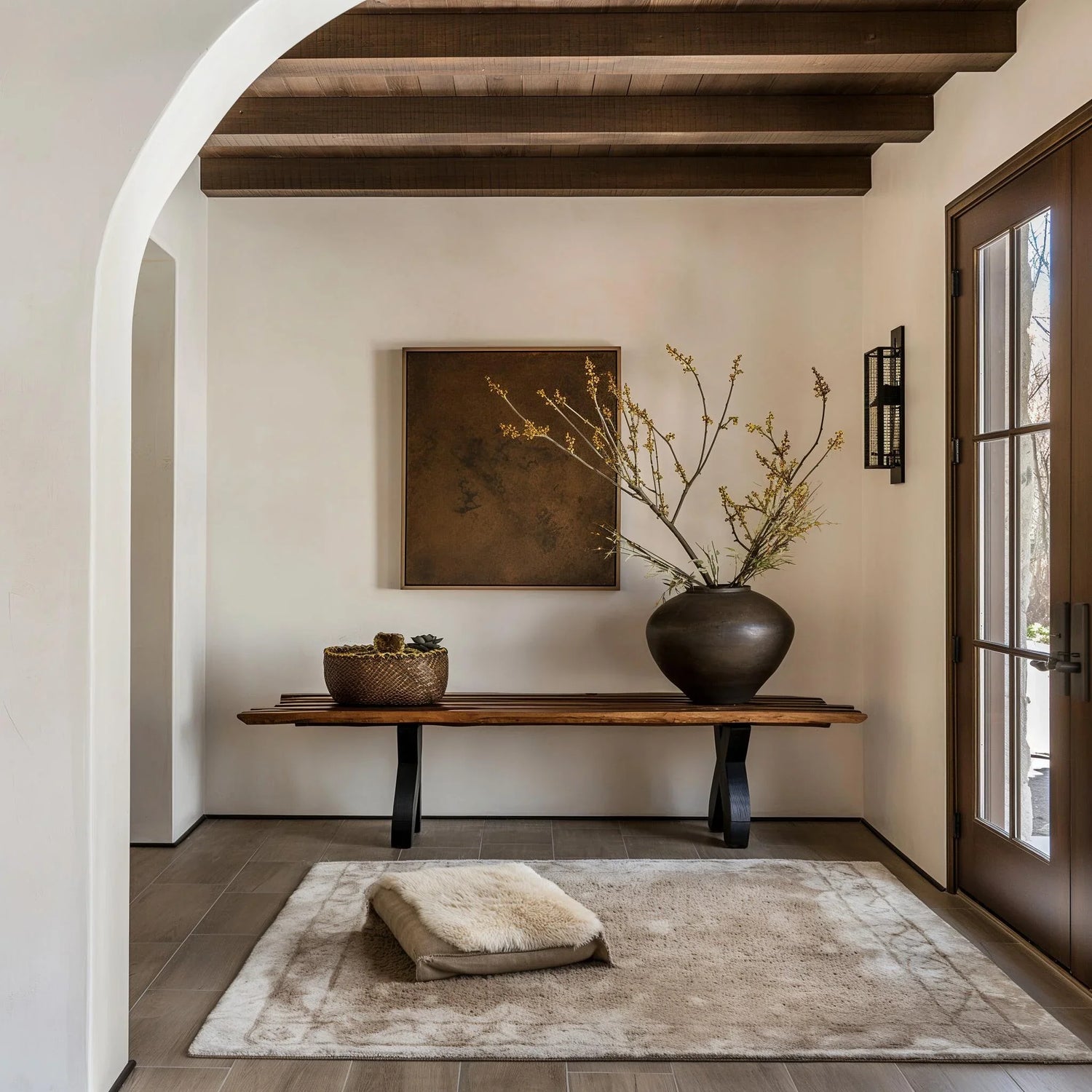 Minimalist entryway with wooden bench, large vase holding dried branches, textured rug, and abstract wall art