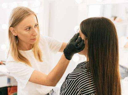 Makeup artist in black gloves applying eyebrow makeup on woman with long brown hair in bright studio