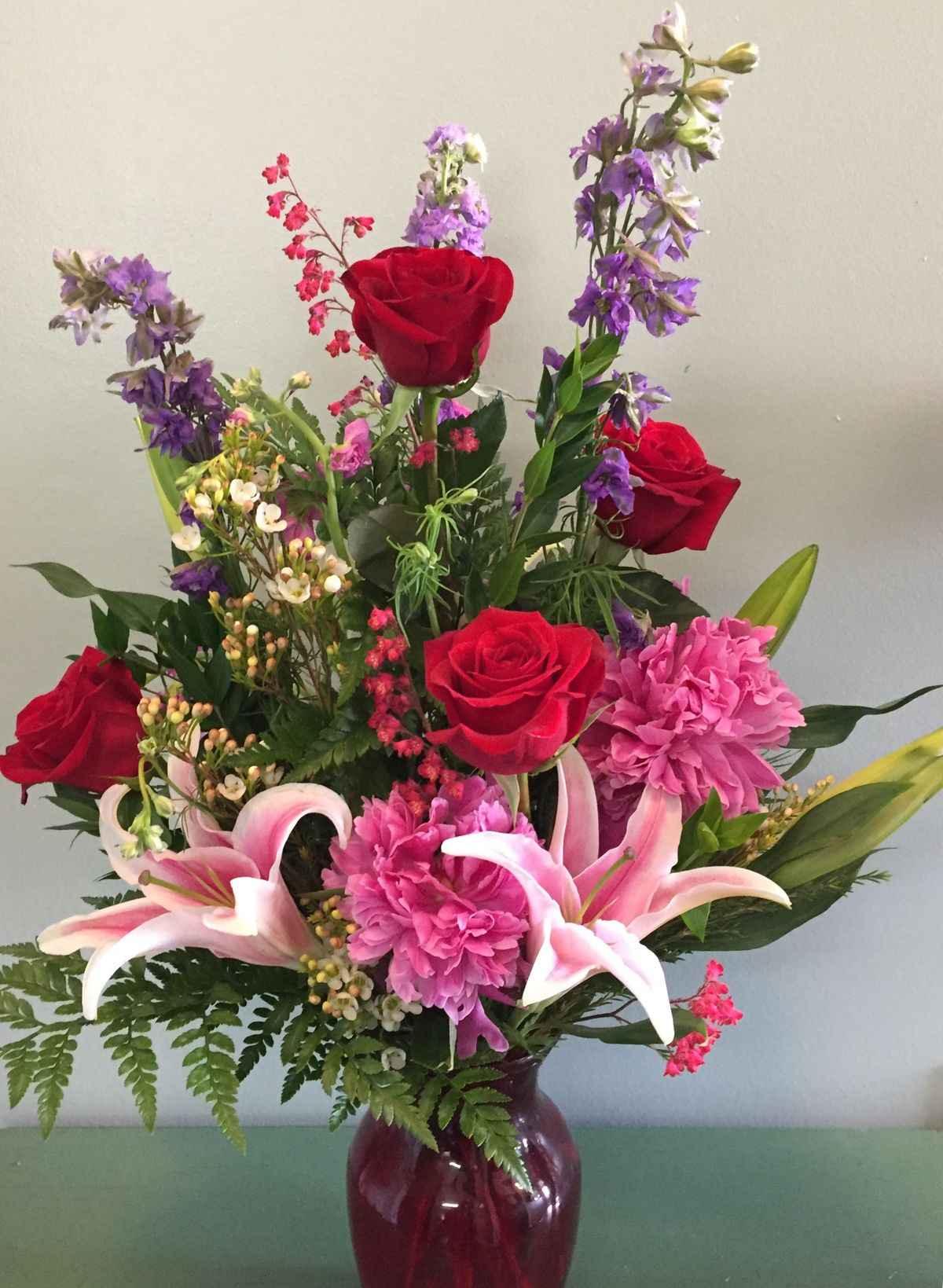 Mixed flower bouquet with red roses, pink lilies, purple flowers, and greenery in a red vase