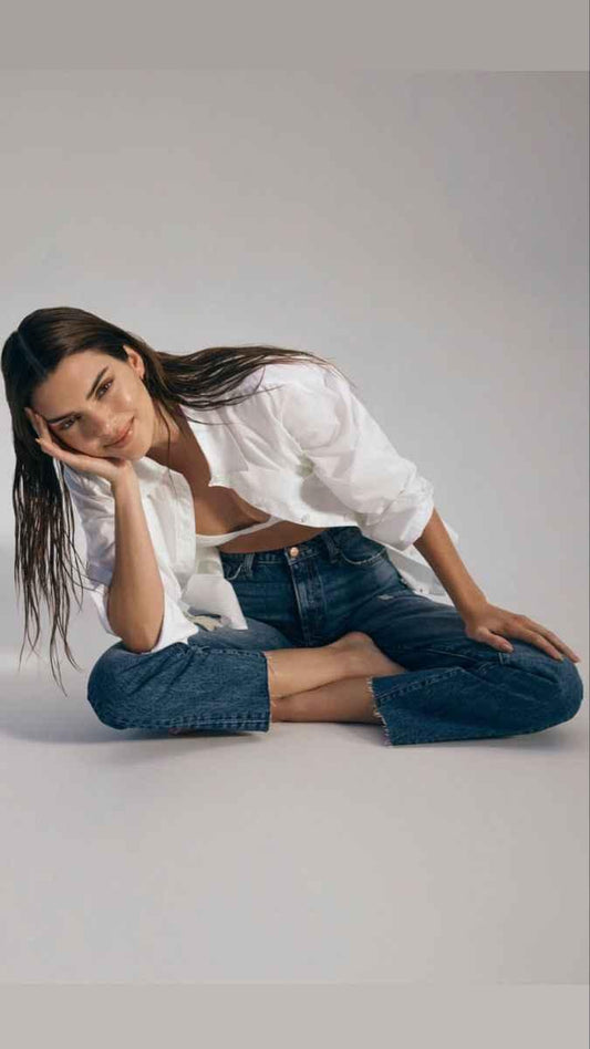 Young woman with long wet hair wearing a white shirt and blue jeans sitting cross-legged on a light background