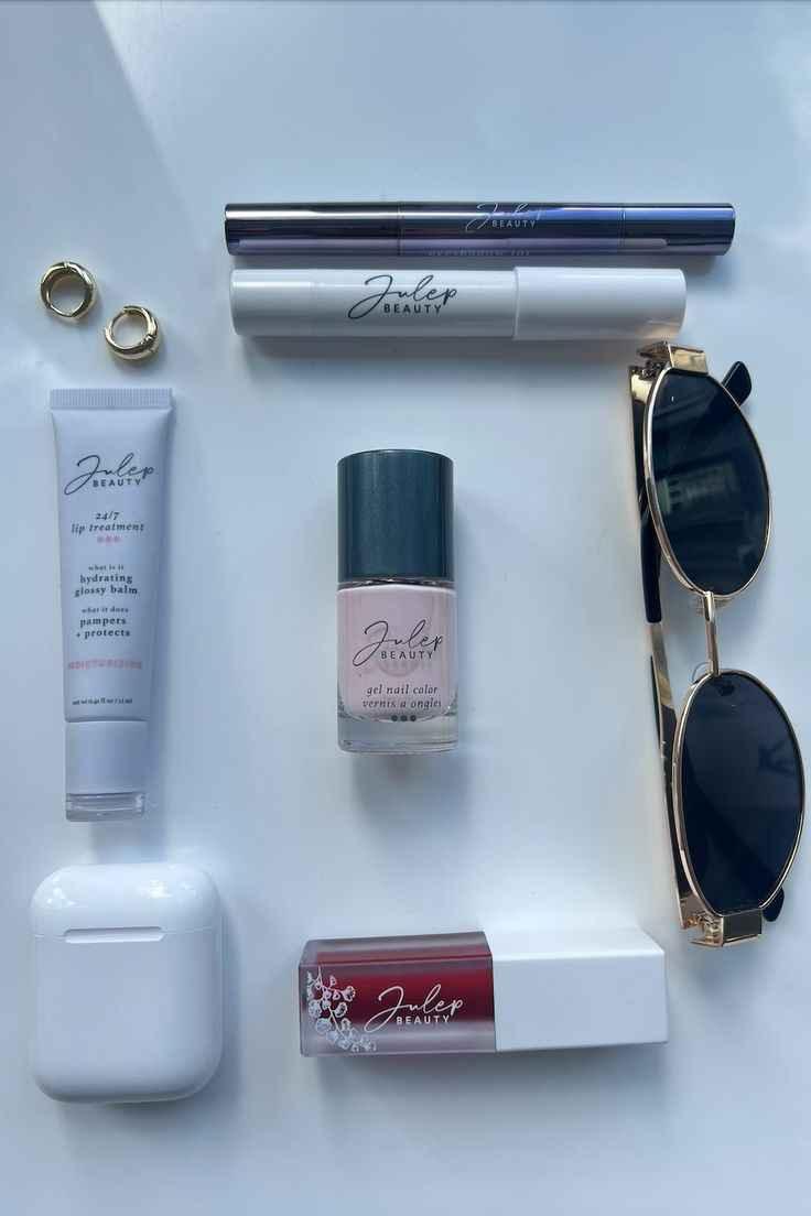 Flat lay of Julep Beauty cosmetics including lip treatment, gel nail color, mascara, lip gloss, gold hoop earrings, sunglasses, and white AirPods case