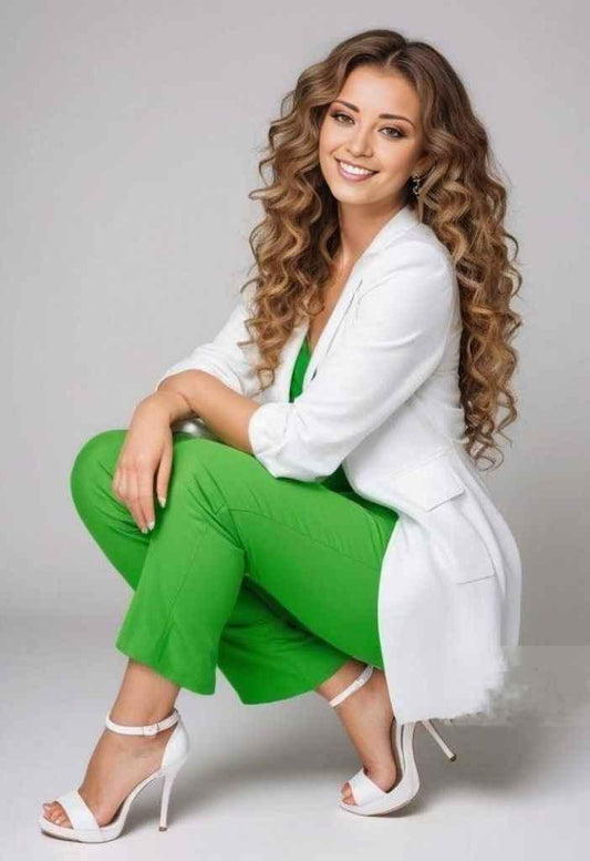 Smiling woman with long curly hair wearing green pants, white blazer, and white heels on gray background