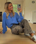 Smiling woman taking a mirror selfie wearing a blue knitted cardigan and leopard print pants indoors