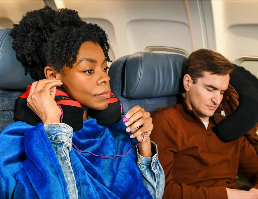 Woman adjusting red and black travel neck pillow and wearing purple earplugs on airplane next to man with brown neck pillow