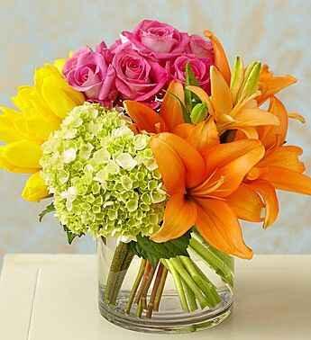 Vibrant floral arrangement with orange lilies, pink roses, yellow tulips, and green hydrangea in a clear glass vase