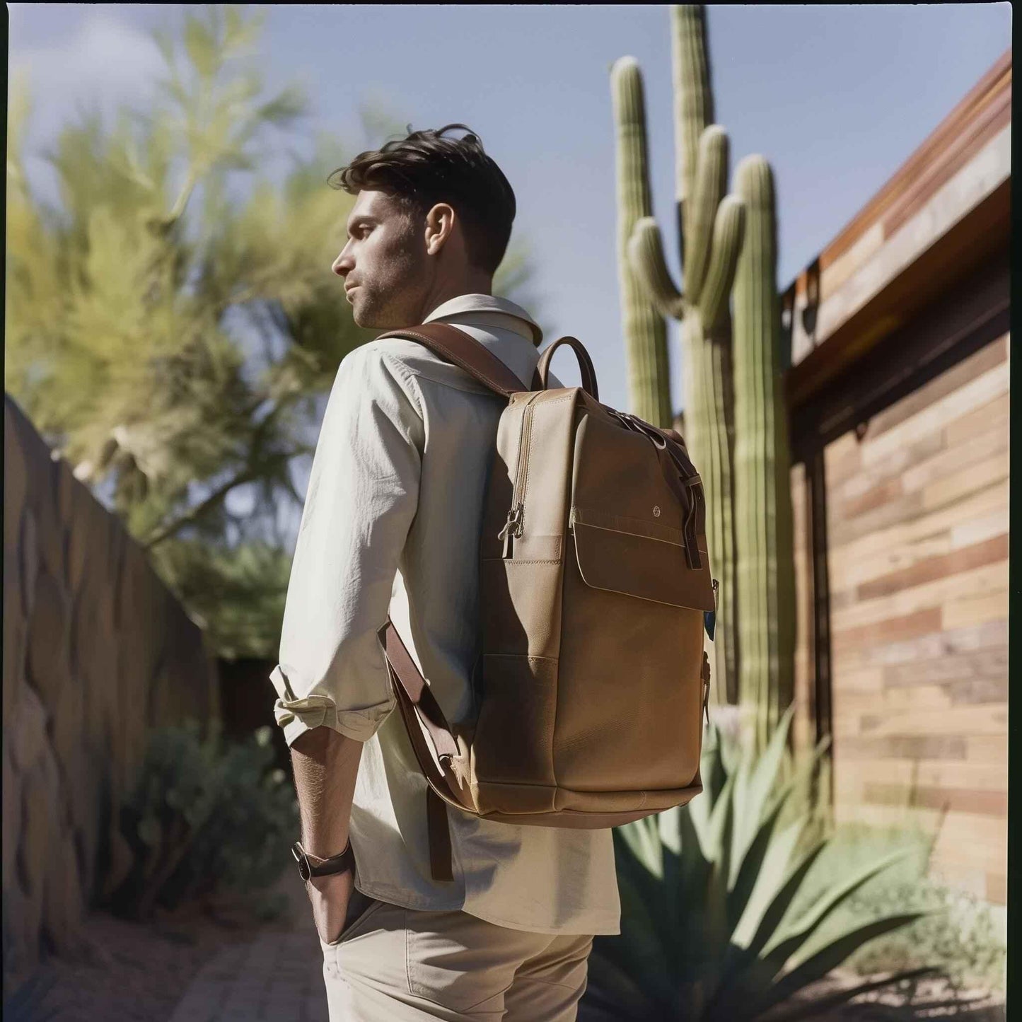 Man in light shirt with stylish tan leather backpack outdoors near cactus and wooden fence